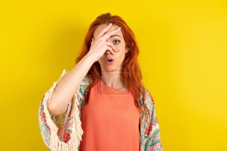 Téléchargez les photos : Femme aux cheveux roux debout sur fond jaune studio regardant sous le choc couvrant le visage et les yeux avec la main, regardant à travers les doigts avec une expression embarrassée. - en image libre de droit