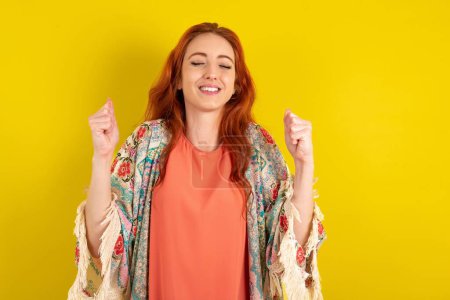 Téléchargez les photos : Femme aux cheveux roux debout sur fond jaune studio étant excité pour le succès avec les bras levés et les yeux fermés célébrant la victoire. Concept gagnant. - en image libre de droit