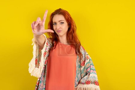 Photo for Red haired woman standing over yellow studio background making fun of people with fingers on forehead doing loser gesture mocking and insulting. - Royalty Free Image