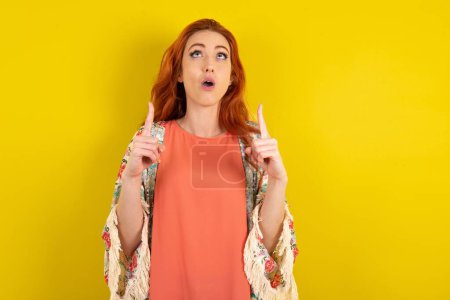 Foto de Mujer pelirroja de pie sobre fondo amarillo estudio asombrado y sorprendido mirando hacia arriba y señalando con los dedos y los brazos levantados. - Imagen libre de derechos