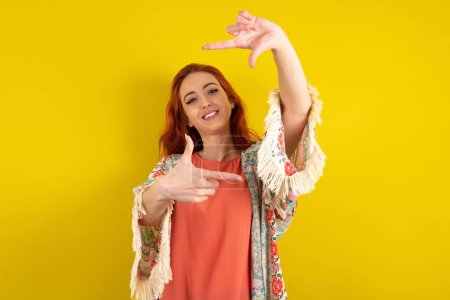 Téléchargez les photos : Femme aux cheveux rouges debout sur fond jaune studio faisant cadre doigt avec les mains. Concept de créativité et de photographie. - en image libre de droit