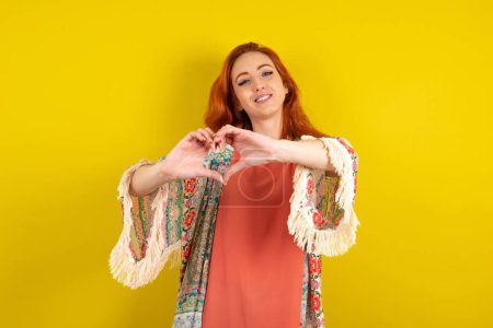 Téléchargez les photos : Femme aux cheveux rouges debout sur fond jaune studio souriant dans l'amour faisant forme de symbole de coeur avec les mains. Concept romantique. - en image libre de droit