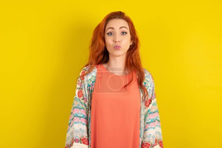 Photo for Shot of pleasant looking red haired woman standing over yellow studio background , pouts lips, looks at camera, Human facial expressions - Royalty Free Image