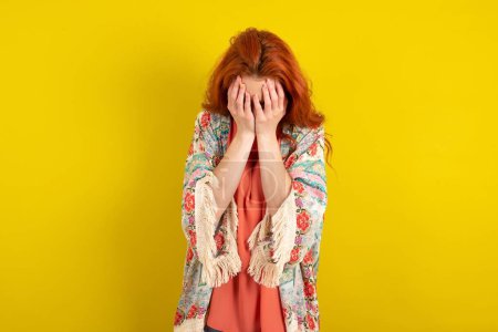 Téléchargez les photos : Femme aux cheveux roux debout sur fond jaune studio couvrant son visage avec ses mains, être dévasté et pleurer. Triste concept - en image libre de droit