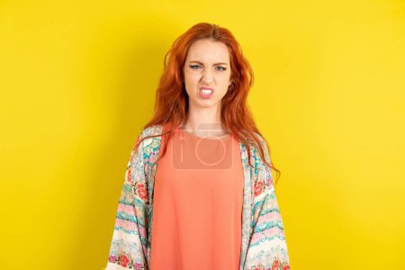 Photo for Red haired woman over yellow studio background keeps teeth clenched, frowns face in dissatisfaction, irritated because of much duties. - Royalty Free Image