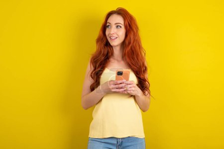 Foto de Hermosa mujer de pelo rojo vistiendo camisa amarilla sobre fondo de estudio amarillo sosteniendo un teléfono inteligente y mirando hacia los lados en blanco copyspace. - Imagen libre de derechos