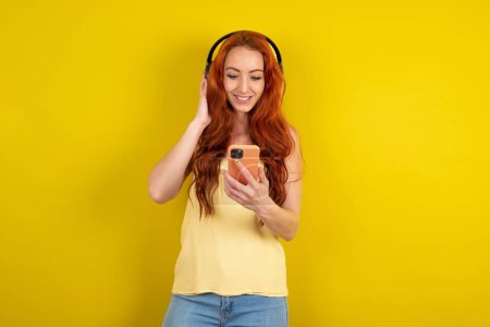 Téléchargez les photos : Heureuse femme aux cheveux rouges portant une chemise jaune sur fond de studio jaune se sent bien tout en se concentrant sur l'écran du smartphone. Personnes, technologie, mode de vie - en image libre de droit