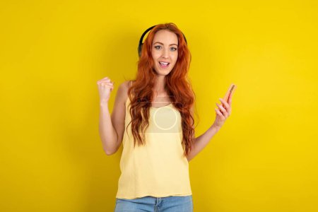 Téléchargez les photos : Belle femme aux cheveux rouges positive portant une chemise jaune sur fond de studio jaune tient un téléphone portable moderne connecté à un casque, serre le poing de bonnes émotions, s'exclame avec joie, - en image libre de droit
