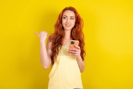 Téléchargez les photos : Belle femme aux cheveux rouges portant chemise jaune sur fond de studio jaune en utilisant et textos avec smartphone pointant et montrant le pouce sur le côté avec sourire heureux visage - en image libre de droit