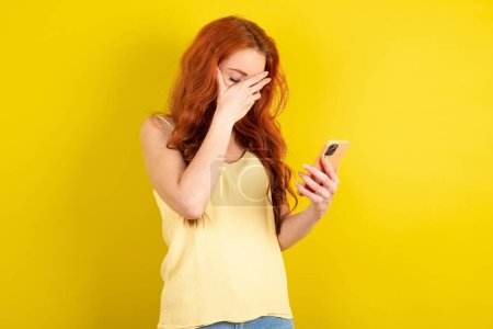 Photo for Beautiful red haired woman wearing yellow shirt over yellow studio background looking at smartphone feeling sad holding hand on face. - Royalty Free Image