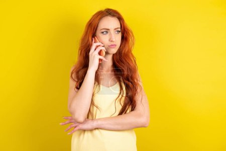 Photo for Sad red haired woman wearing yellow shirt over yellow studio background talking on smartphone. Communication concept. - Royalty Free Image