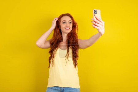 Téléchargez les photos : Belle femme aux cheveux rouges portant une chemise jaune sur fond de studio jaune souriant et prenant un selfie prêt à le poster sur ses médias sociaux. - en image libre de droit