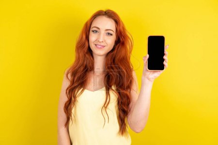 Téléchargez les photos : Belle femme aux cheveux rouges souriante portant une chemise jaune sur fond de studio jaune avec un espace de copie simulé. Maintenez le téléphone portable avec écran vide vide - en image libre de droit