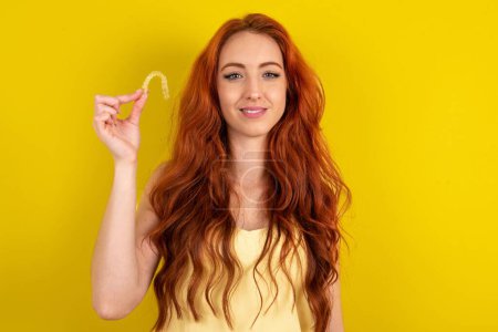 Photo for Beautiful red haired woman wearing yellow shirt over yellow studio background holding an invisible braces aligner, recommending this new treatment. Dental healthcare concept. - Royalty Free Image
