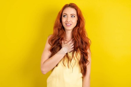 Photo for Joyful beautiful red haired woman wearing yellow shirt over yellow studio background expresses positive emotions recalls something funny keeps hand on chest and giggles happily. - Royalty Free Image