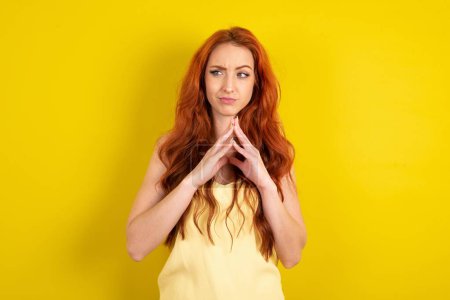 Téléchargez les photos : Belle femme aux cheveux rouges portant chemise jaune sur fond jaune studio doigts raides et semble mystérieux de côté a grand plan maléfique à l'esprit - en image libre de droit