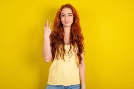 Foto de Hermosa mujer de pelo rojo vistiendo camisa amarilla sobre fondo amarillo estudio sonriendo y buscando amigable, mostrando el número tres o tercero con la mano hacia adelante, cuenta atrás - Imagen libre de derechos