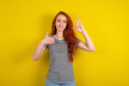 Foto de Mujer pelirroja vistiendo camisa a rayas sobre fondo amarillo estudio sonriendo y luciendo feliz, despreocupada y positiva, haciendo gestos de victoria o paz con una mano - Imagen libre de derechos