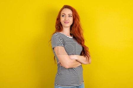 Foto de Mujer de pelo rojo vistiendo camisa a rayas sobre los brazos de fondo de estudio amarillo cruzados mirar cartelera espacio vacío - Imagen libre de derechos