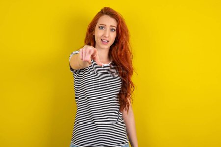 Foto de Emocionada mujer de pelo rojo positivo con camisa a rayas sobre los puntos de fondo del estudio amarillo dedo índice directamente en usted, ve algo muy divertido. Wow, increíble - Imagen libre de derechos