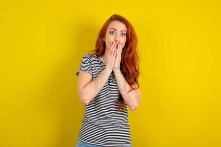 Photo for Red haired woman wearing striped shirt over yellow studio background keeps hands on mouth, looks with eyes full of disbelief, being puzzled with amount of work - Royalty Free Image