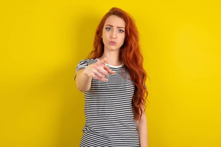 Photo for Shocked red haired woman wearing striped shirt over yellow studio background points at you with stunned expression - Royalty Free Image