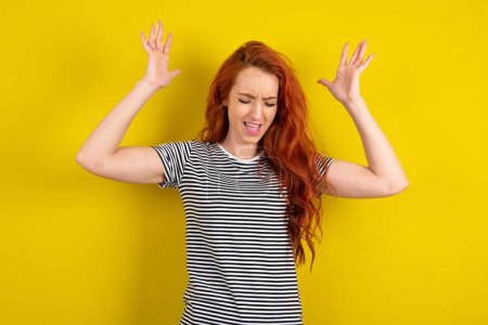 Téléchargez les photos : Femme aux cheveux rouges portant une chemise rayée sur fond jaune studio devient fou comme la tête tourne se sent stressé en raison de la situation horrible - en image libre de droit