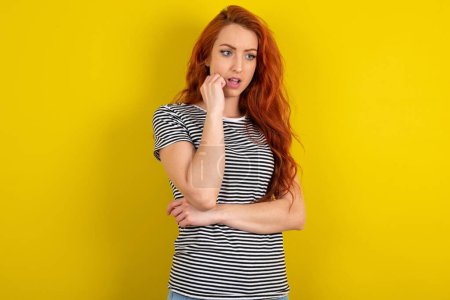 Photo for Astonished red haired woman wearing striped shirt over yellow studio background looks aside surprisingly with opened mouth. - Royalty Free Image