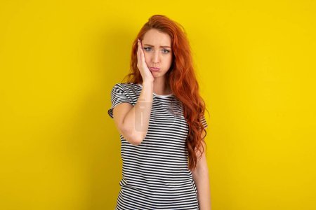 Photo for Sad lonely red haired woman wearing striped shirt over yellow studio background touches cheek with hand bites lower lip and gazes with displeasure. Bad emotions - Royalty Free Image