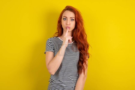 Photo for Surprised red haired woman wearing striped shirt over yellow studio background makes silence gesture, keeps finger over lips and looks mysterious at camera - Royalty Free Image