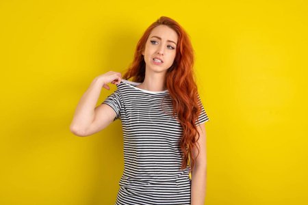 Téléchargez les photos : Femme aux cheveux rouges portant une chemise rayée sur fond jaune studio stressé, anxieux, fatigué et frustré, tirant col de chemise, l'air frustré par le problème - en image libre de droit