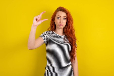 Photo for Red haired woman wearing striped shirt over yellow studio background purses lip and gestures with hand, shows something very little. - Royalty Free Image