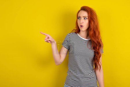 Photo for Stunned red haired woman wearing striped shirt over yellow studio background with greatly surprised expression points away on copy space, indicates something - Royalty Free Image