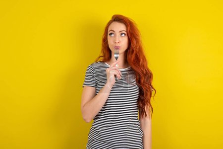 Photo for Photo of dreamy red haired woman wearing striped shirt over yellow studio background lick fork look, empty space - Royalty Free Image
