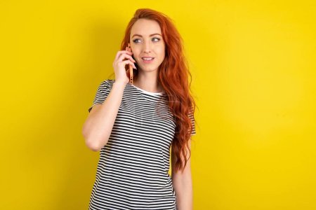 Photo for Pleasant looking happy red haired woman wearing striped shirt over yellow studio background has nice telephone conversation and looks aside, has nice mood and smiles positively while talks via cell phone - Royalty Free Image