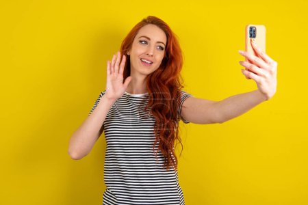 Foto de Mujer de pelo rojo con camisa a rayas sobre fondo de estudio amarillo sostiene teléfono móvil moderno y hace que las ondas de videollamada palma en gesto de hola. Concepto de tecnología moderna - Imagen libre de derechos