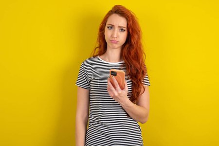 Foto de Mujer cabellera roja insatisfecha con camisa rayada sobre fondo de estudio amarillo utiliza la aplicación de software móvil y la información de navegación en Internet, sostiene la mano móvil moderna - Imagen libre de derechos
