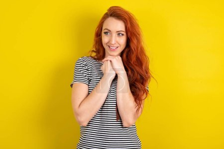 Photo for Happy red haired woman wearing striped shirt over yellow studio background anticipates something awesome happen, looks happily aside, keeps hands together near face, has glad expression. - Royalty Free Image