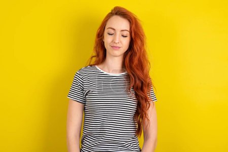 Téléchargez les photos : Femme aux cheveux rouges portant une chemise rayée sur fond de studio jaune beau doux charme mignon attrayant beau winsome doux yeux fermés paisibles - en image libre de droit