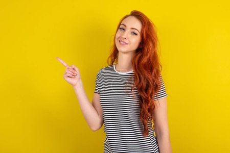 Photo for Profile photo of red haired woman wearing striped shirt over yellow studio background indicate finger with empty space offer - Royalty Free Image