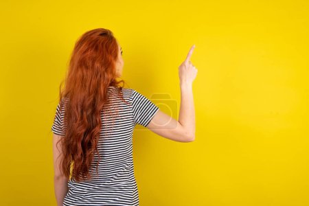 Téléchargez les photos : Femme aux cheveux rouges portant une chemise rayée sur fond jaune studio pointant vers l'objet sur l'espace de copie, vue arrière. Tourne le dos - en image libre de droit