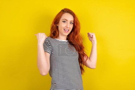 Téléchargez les photos : Cool femme aux cheveux rouges portant une chemise rayée sur fond jaune studio point de retour sur l'espace vide avec poing à la main - en image libre de droit