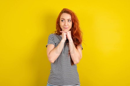 Photo for Curious red haired woman wearing striped shirt over yellow studio background keeps hands under chin bites lips and looks with interest aside. - Royalty Free Image