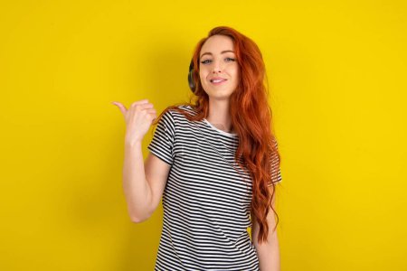 Téléchargez les photos : Femme aux cheveux rouges portant une chemise rayée sur fond jaune studio écoute la piste audio via des écouteurs sans fil points pouce loin annonces espace de copie - en image libre de droit