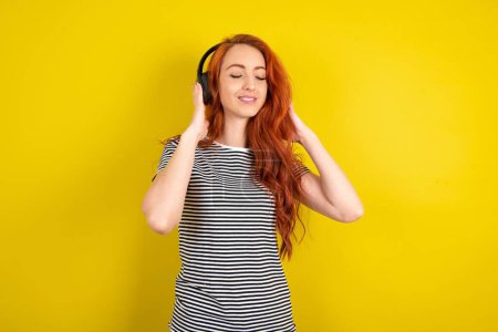 Téléchargez les photos : Femme aux cheveux rouges portant une chemise rayée sur fond de studio jaune avec un casque sur la tête, écoutant de la musique, appréciant la chanson préférée avec les yeux fermés, tenant la main sur un casque. - en image libre de droit