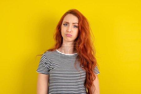 Photo for Displeased red haired woman wearing striped shirt over yellow studio background frowns face feels unhappy has some problems. Negative emotions and feelings concept - Royalty Free Image