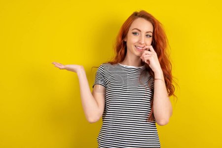 Photo for Positive red haired woman wearing striped shirt over yellow studio background  advert promo when touching teeth with finger - Royalty Free Image