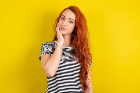Téléchargez les photos : Photo de femme aux cheveux rouges portant une chemise rayée sur fond jaune studio profiter d'une peau fraîche et lisse toucher visage - en image libre de droit