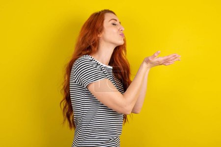 Téléchargez les photos : Profil vue de côté, portrait d'une jolie femme aux cheveux rouges portant une chemise rayée sur fond jaune studio envoyant un baiser d'air - en image libre de droit