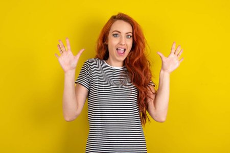 Foto de Deliciosa mujer de pelo rojo positivo con camisa a rayas sobre fondo de estudio amarillo abre la boca y los brazos palmeras después de tener un gran resultado - Imagen libre de derechos